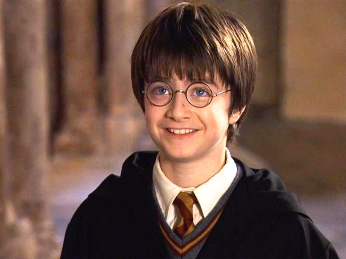 Harry Potter And The Philosopher`s Stone / Harry Potter Felsefe Taşı Filmi - Harry Potter Film içerisinden bir kare olan yatay dikdörtgen fotoğrafta göğsünden yukarısı gözüken Harry Potter (Daniel Radcliffe) yukarıya doğru bakarak gülümsüyor. Arka planda kahverengi tonlarının ağırlıkta olduğu bir alan (salon) var.  Harry Potter 11 yaşında bir oğlan çocuğu. Beyaz bir ten rengine sahip. Yuvarlak ve keskin olmayan yüz hatları var. Düz ve kahverengi saçları alnına düşüyor. Yandan uzanan saçları ise kulaklarının yarısında son buluyor.  Kaşları saçlarının arkasında kalmış. Mavi ve büyük gözleri hafif kısılmış. Siyah çerçeveli yuvarlak gözlükleri var.  Gülümsemesi sebebiyle burun kenarlarından dudak çevresine doğru çizgiler belirmiş ve yanakları ön plana çıkmış. Üst dudağı alt dudağından daha ince ve sadece büyük ve beyaz ön dişleri gözüküyor. İnce kemerli ama geniş kanatlı bir burnu var. Burnunun sağ yanında küçük kırmızı bir yara var. Harry’nin üstünde siyah okul cüppesi var. Beyaz renkte gömleğinin üzerine sarı ve kırmızı şeritleri olan bir kravat bağlamış. Gömlek ve kravatın üstüne V yakalı ve yakasında kırmızı sarı şerit bulunan gri renkte bir kazak giymiş.