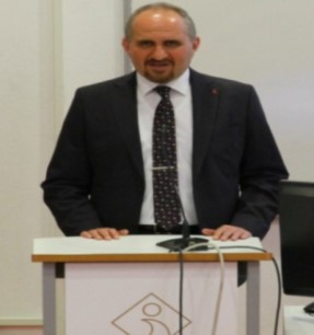Sesli Betimlemeyle Yeni Bir Dünyanın Kapıları Aralanıyor Olgun Yılmaz bir konuşma kürsüsünde konuşuyor. Yılmaz siyah bir takım elbise giymiş.